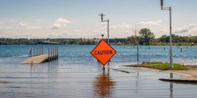 Rising sea water partially submerges a footbridge and has flooded a nearby road. There is an orange caution sign in the foreground.