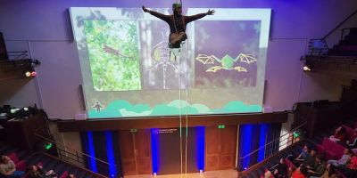 Robin Hayward at the top of a lecture theatre hanging by a rope. Behind them is a screen showing animals from the rainforest, and below are audience members in the theatre.